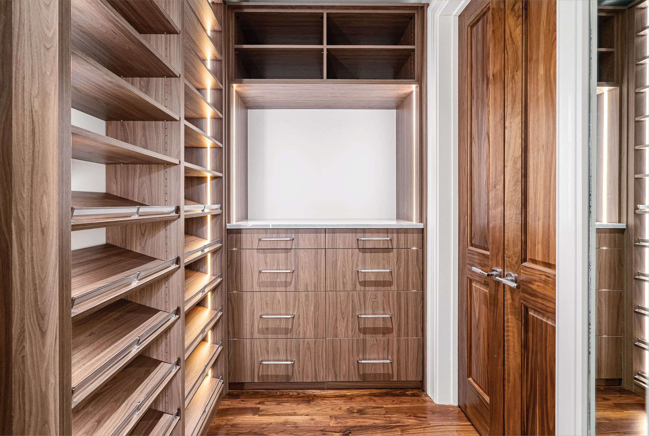 Walk-in primary closet fitted in walnut with lit shelving and built-in drawers